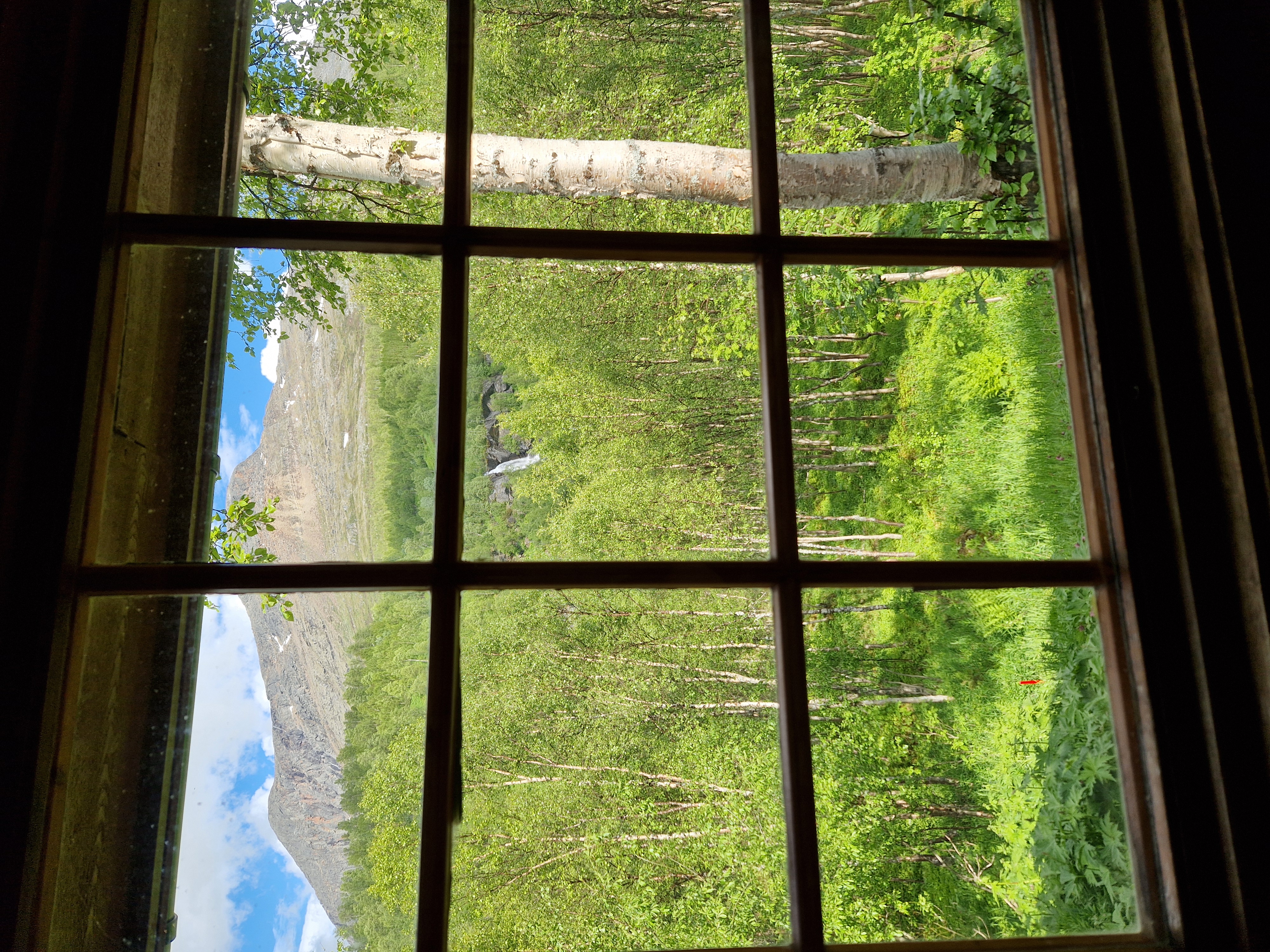 Utsikt ut av et hyttevindu. Det er bjørkeskog, et høyt fjell og en foss utenfor vinduet.